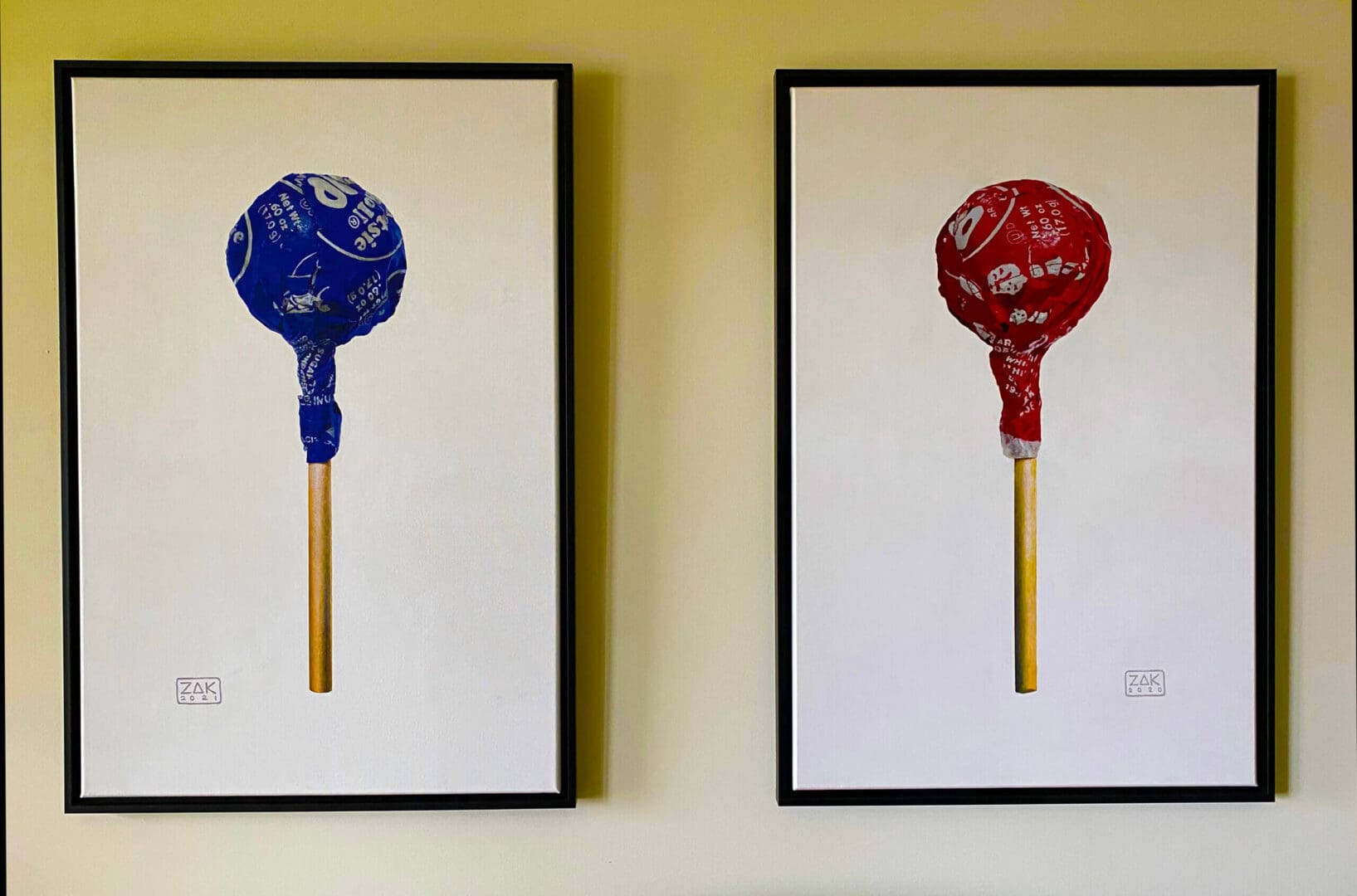 Two framed artworks of lollipops, one blue and one red, on a yellow wall.